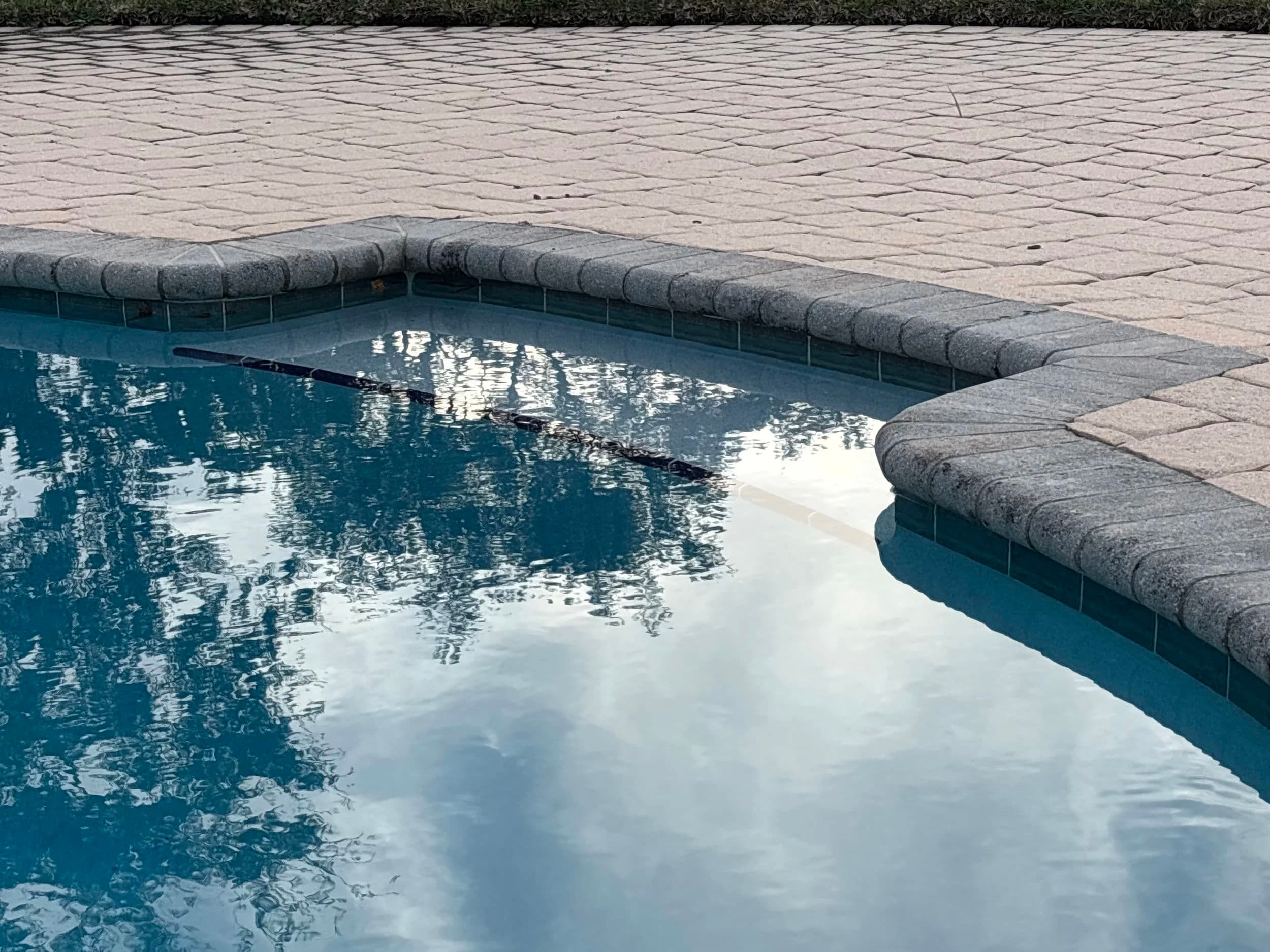 Calm swimming pool reflecting clouds and trees, with a stone edge and clean water.